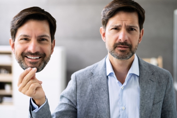 A white man with short dark hair, moustache and stubble and wearing a grey jacket, looks morose while holding up a life sized cut out photograph of his face in which he is smiling. The background is an office with a cabinet.